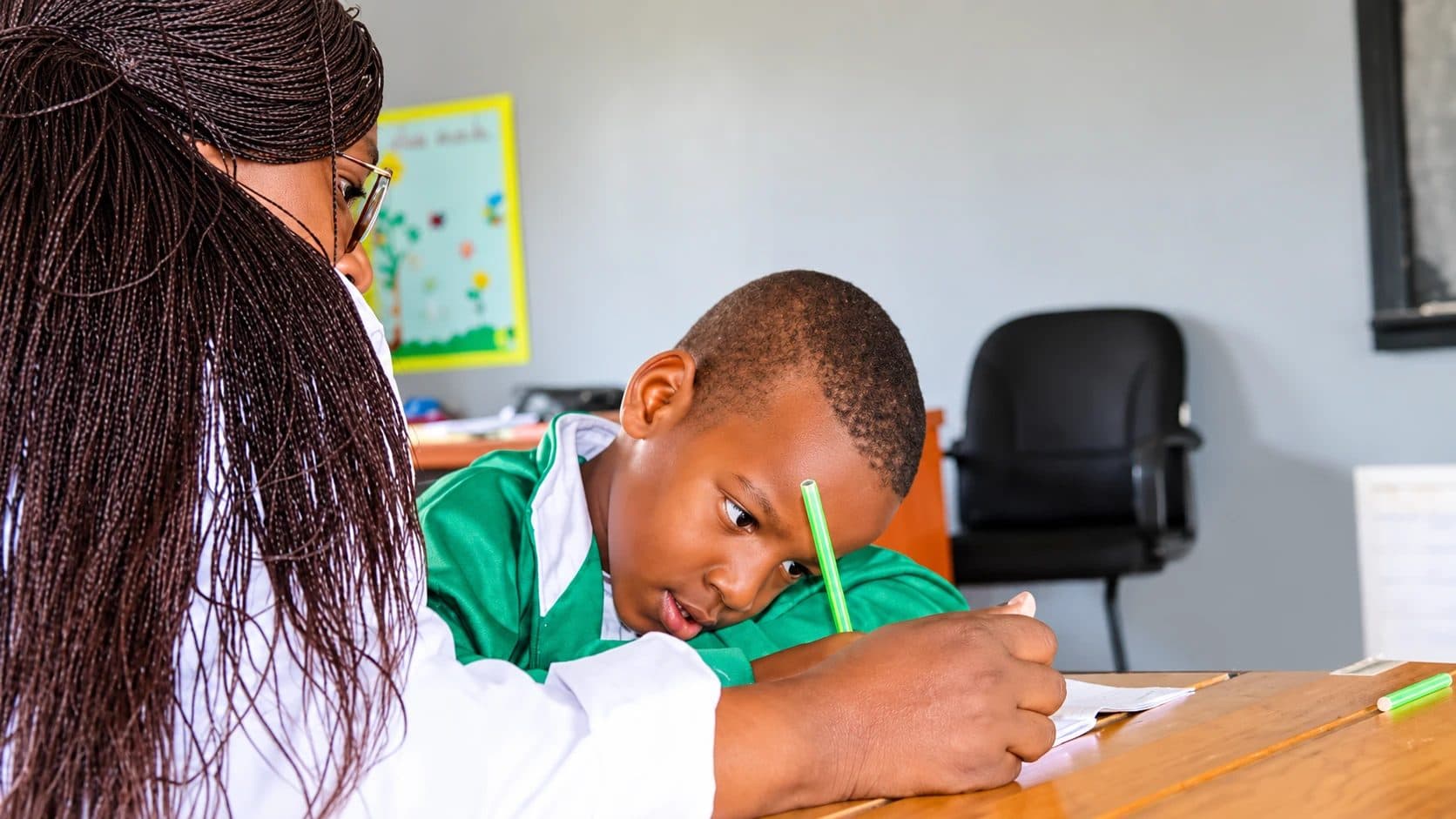 A teacher guiding a young pupil in a green tracksuit as he writes at his desk