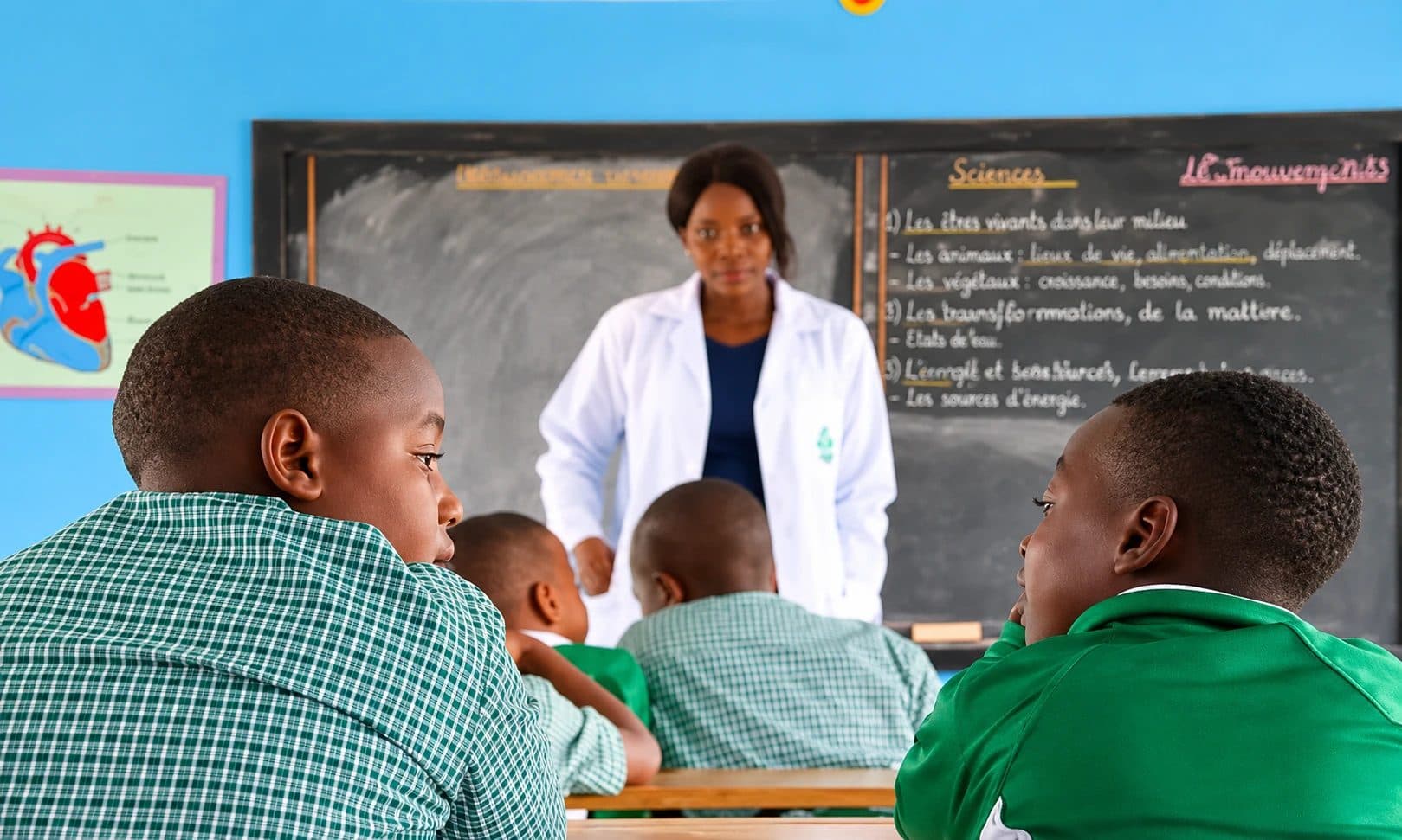 A teacher in a white coat leading a science lesson at the chalkboard while pupils in green uniforms listen