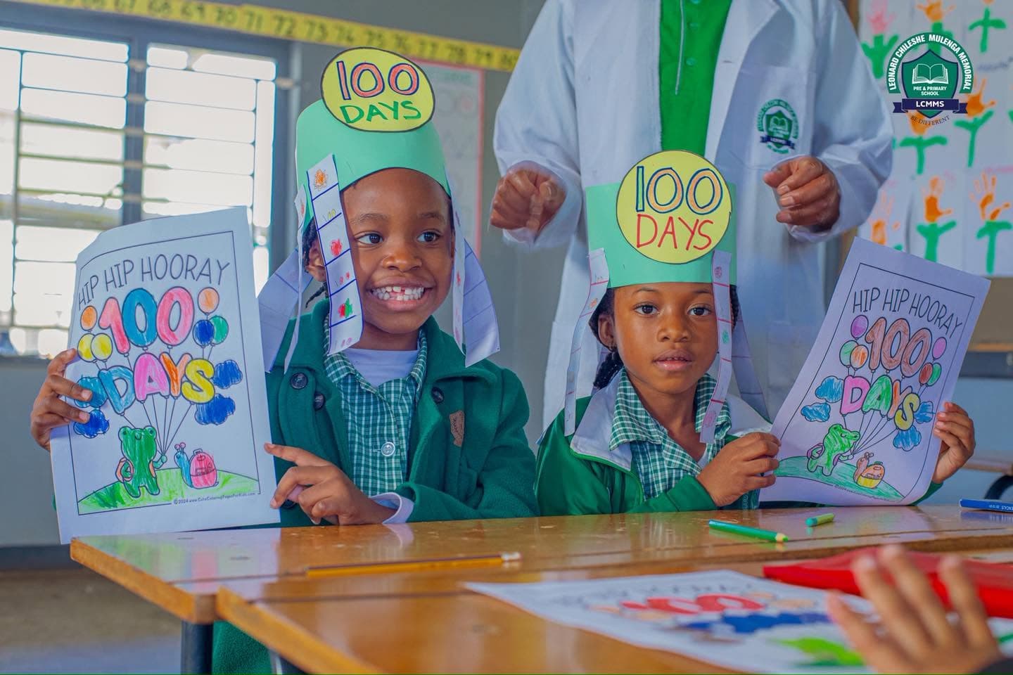 Two learners wearing handmade 100 Days crowns hold up their colourful celebration worksheets