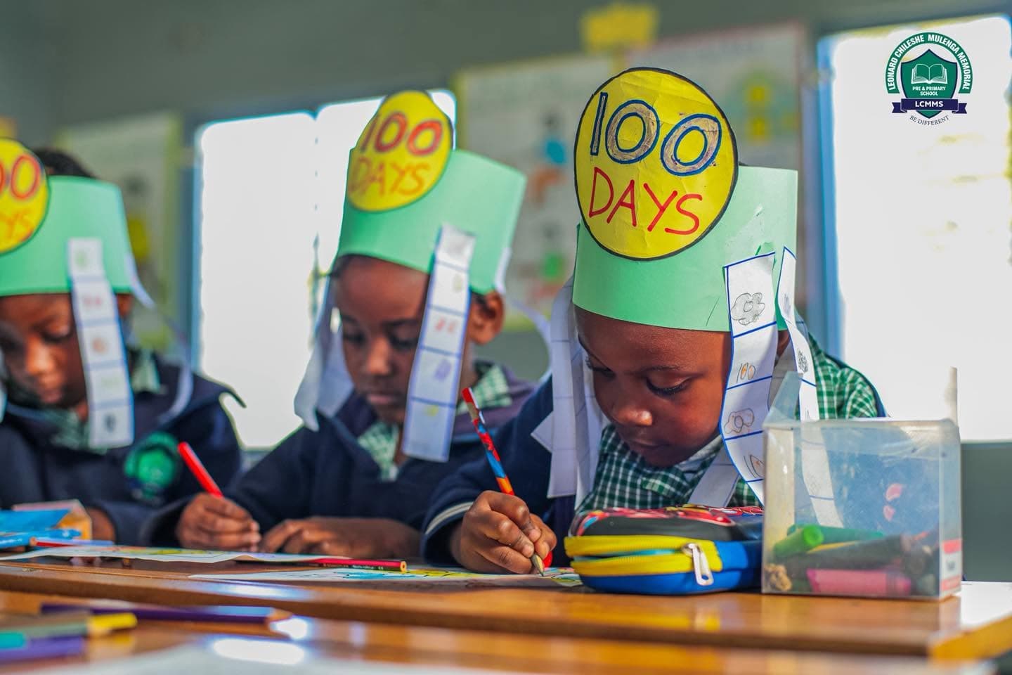 Learners wearing green paper crowns concentrate on colouring at their desks