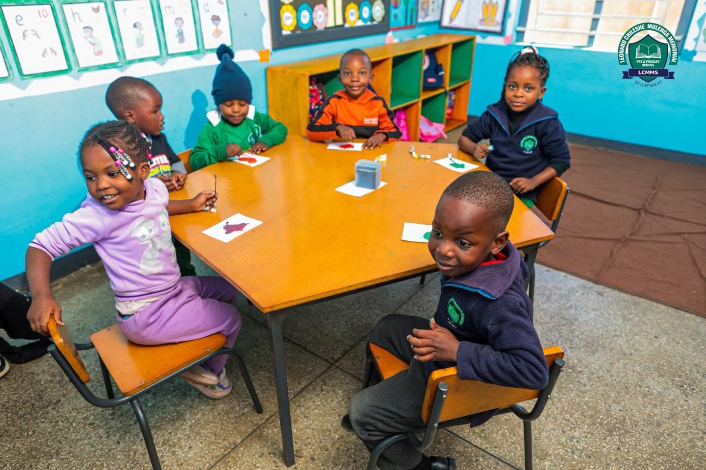 Children sit together around a classroom table with their artwork