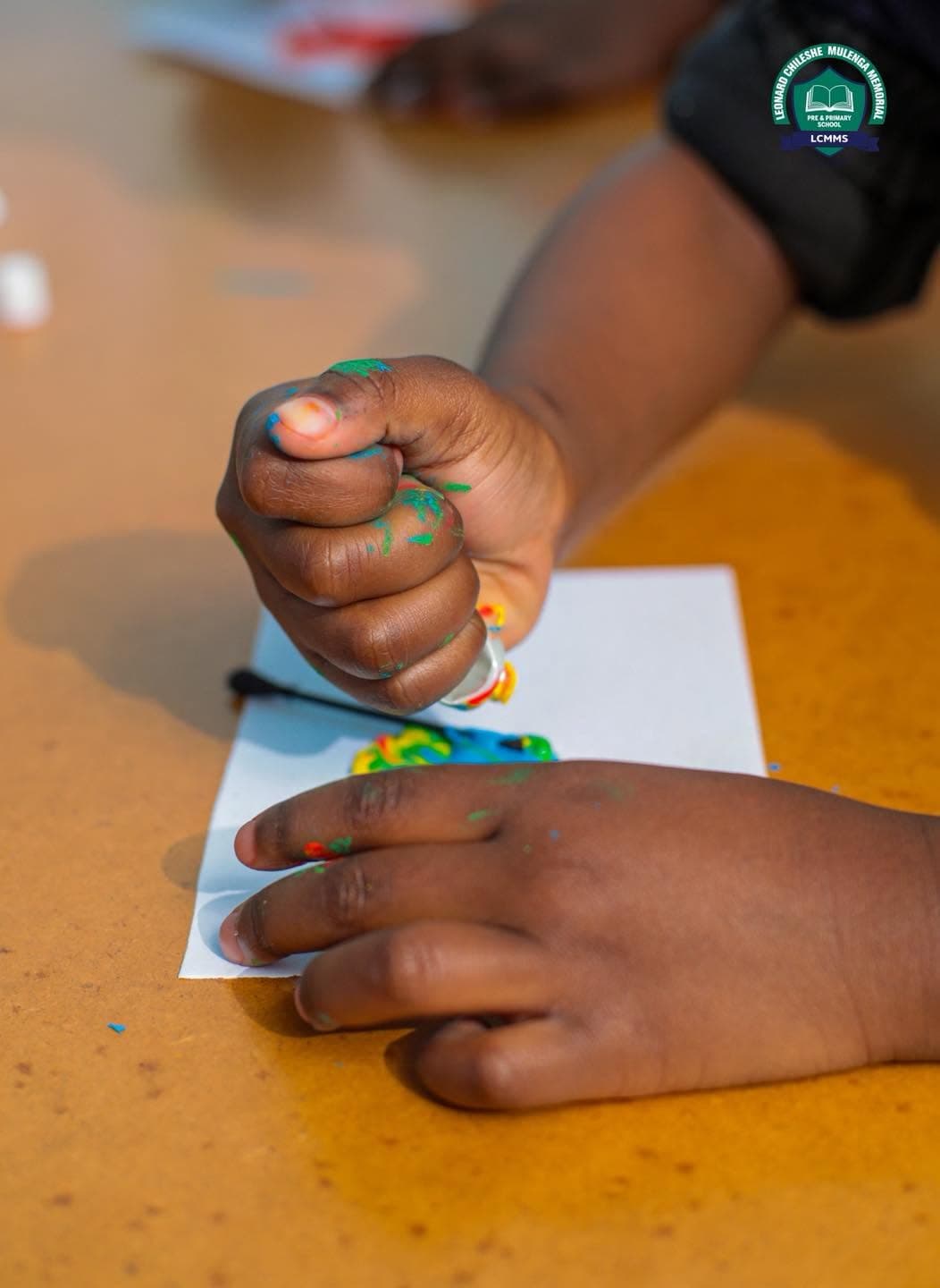 A child's paint-covered hands work on a colourful classroom art activity