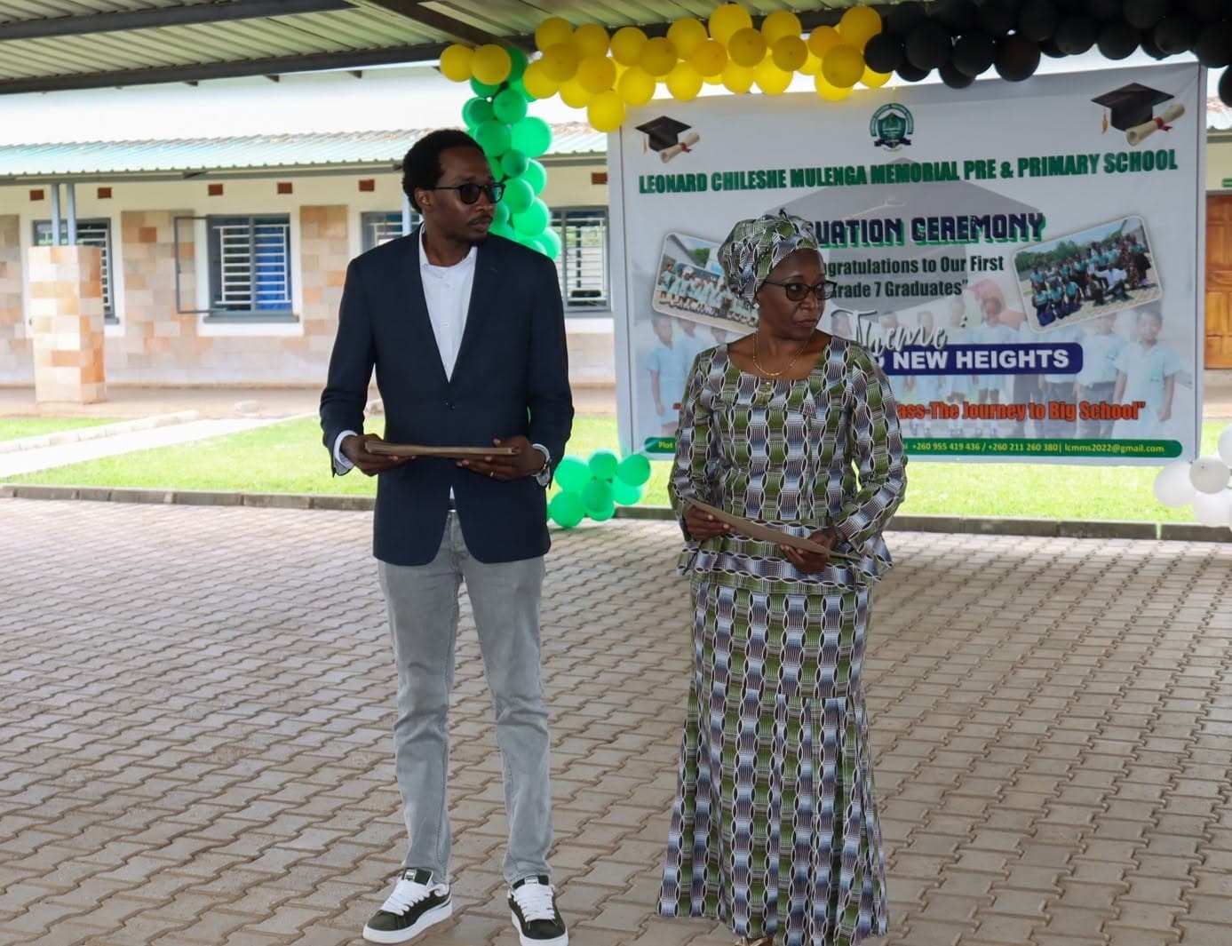 Two adults stand beside the graduation ceremony banner in the decorated school courtyard