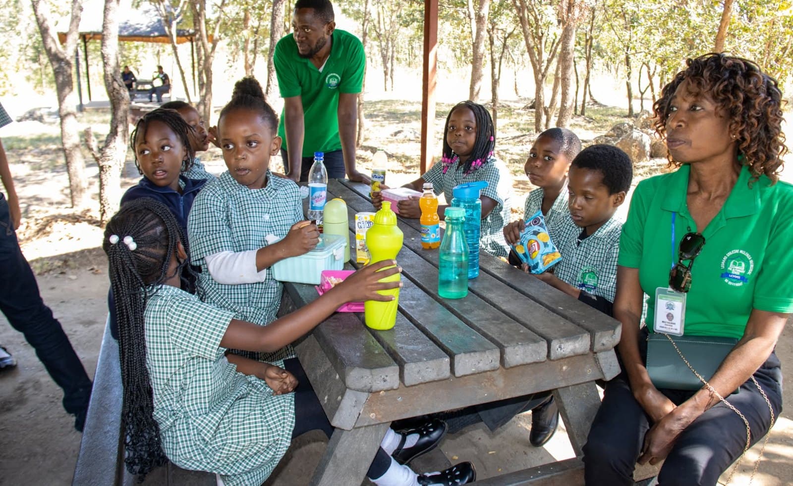Learners and a staff member sit at a shaded picnic table during the school outing