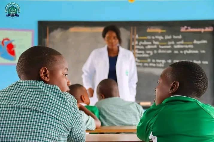 A teacher leading a lesson at the chalkboard for pupils in green uniforms