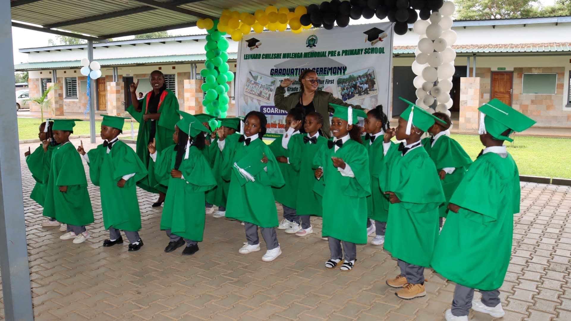 Pre-school graduates in green caps and gowns dancing with their teachers at the school graduation ceremony