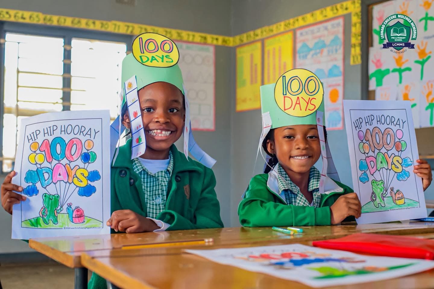 Two smiling learners display their 100 Days artwork at a classroom desk