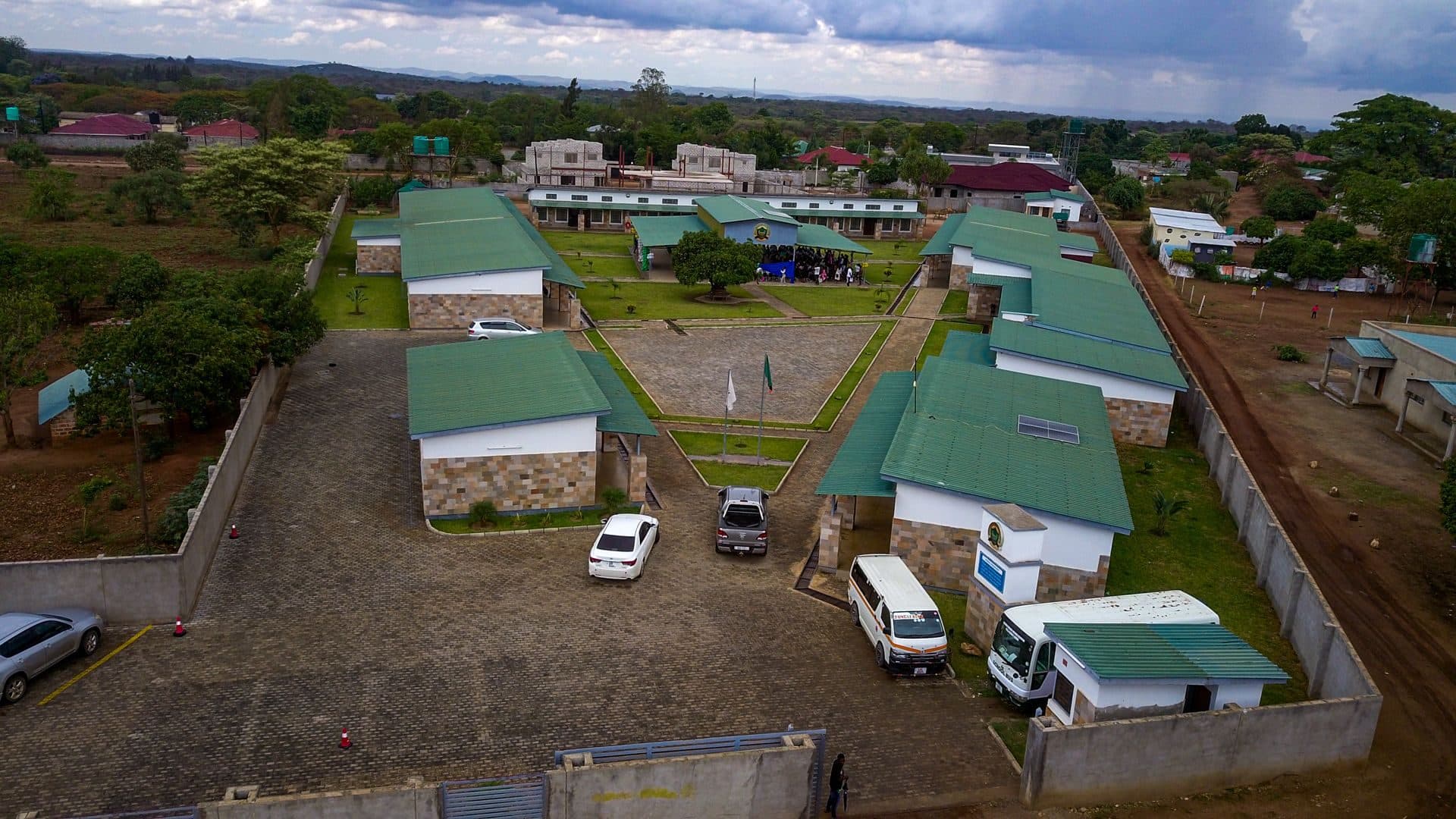 Aerial view of the LCMMS campus: green-roofed classroom blocks around a paved courtyard, with the school bus at the entrance
